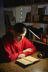 Young man is reading an old book in the library under a table lamp