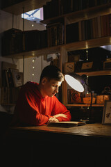 Young man is reading an old book in the library under a table lamp