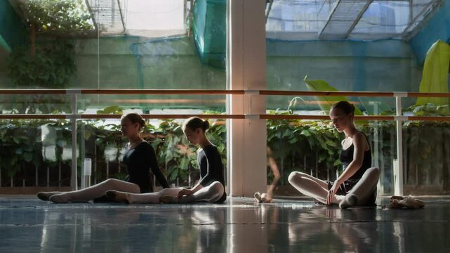 Three young female ballet dancers in black leotards doing stretching exercises on parquet floor in modern dance studio with floor-to-ceiling windows and soft warm light