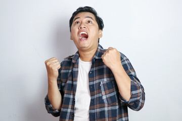 A young Asian man in a casual shirt pumps his fists in excitement and smiles broadly against a solid yellow background. This image captures the essence of celebration and triumph