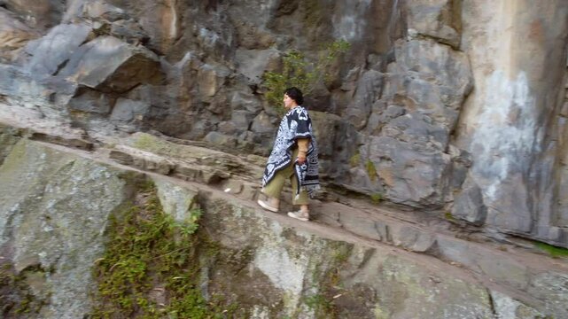 A woman wearing a traditional poncho walks on a narrow mountain path beside a rocky cliff