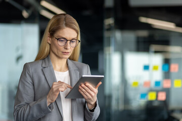 Confident businesswoman in gray suit using digital tablet in bright modern office. She looks focused, embodying professionalism and technology-driven work environment.