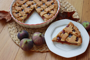 Slice of tart with dark fig jam on a white dessert plate  on wooden table with fresh figs and autumnal leaves