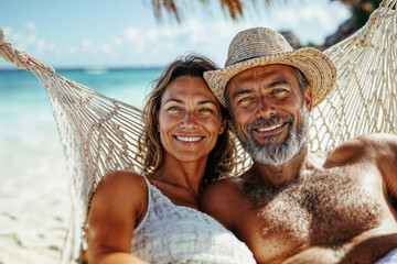 A mature couple is sitting in a hammock on a beach, smiling and enjoying the sun on their vacation.