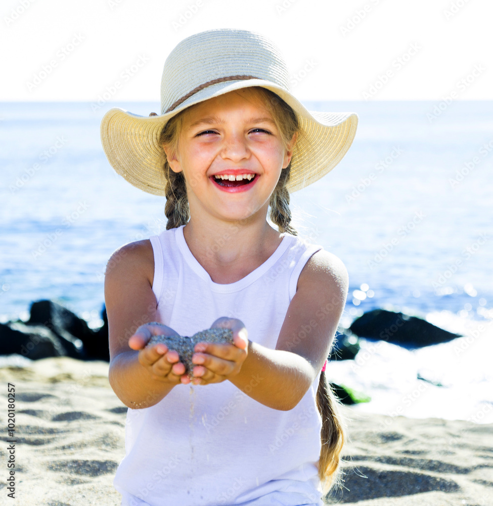 Wall mural Baby girl in hat playing with sand on sea coast in summer