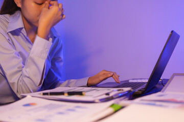 A young woman works overtime at night at her desk in office, using a laptop reviewing documents. is...