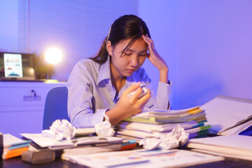 A young woman works overtime at night at her desk in office, using a laptop reviewing documents. is...