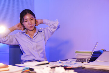 A young woman works overtime at night at her desk in office, using a laptop reviewing documents. is stressed, tired,experiencing headaches,body aches from sitting too long,leading to office syndrome