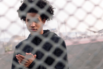 portrait of thoughtful teen african american male with curly hair standing behind metal chain link...