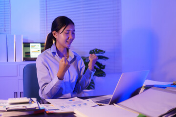 An Asian woman is smiling happily at her desk in the office, having successfully submitted a...