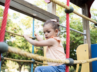 A young girl with disability climbs a set of colorful ropes at a playground, surrounded by trees on a bright, sunny day.