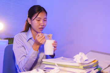 An Asian woman works late at her desk in the office, eating instant noodles while looking at documents and working on her laptop. She is busy with overtime submissions and business tasks at night