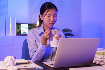 An Asian woman works late at her desk in the office, eating instant noodles while looking at documents and working on her laptop. She is busy with overtime submissions and business tasks at night