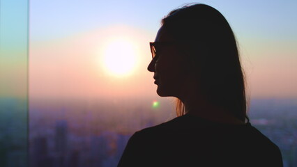 Woman enjoy aerial sunset city skyline standing near the window. Long hair girl silhouette in sunglasses against bright colorful sky, sun goes down.