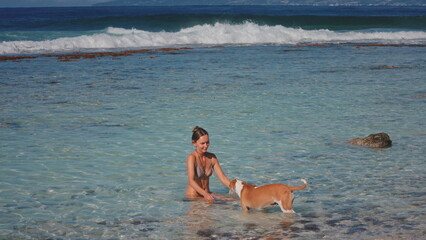 A woman is sitting in the water next to a dog. The woman appears relaxed she pets the dog gently....
