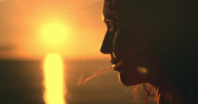 Close up woman face against bright orange sunset. Girl silhouette enjoys amazing colorful sunset, standing on a tropical beach, raises hands against orange dramatic sky and ocean waves. Holiday travel
