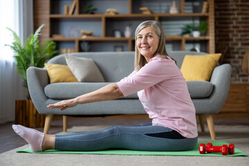 Smiling mature woman doing yoga stretch on mat indoors. Wearing comfortable activewear, she enjoys...