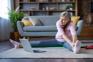 Mature woman stretches on yoga mat with laptop guidance, exemplifying active lifestyle at home. Senior practicing yoga for health and flexibility in living room setting, embracing wellness.