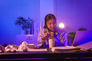 Young Asian business trainee works late at her desk, surrounded by papers and a laptop. Overtime stress builds as she eats noodles, trying to stay focused while battling exhaustion and deadlines.