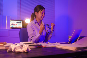 young Asian business trainee works late at her desk, typing on a laptop. Despite the overtime, she...
