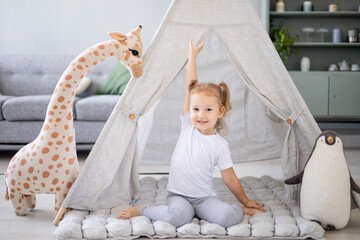 a blonde girl in a bright children's room plays wooden cubes with a giraffe at the wigwam and rejoices and smiles, raising her hands up © Any Grant