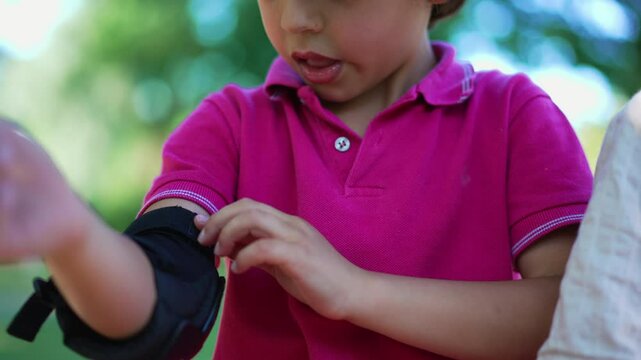 Young boy fastening elbow pad, focused on adjusting protective gear before playing, showing concentration and attention to safety for outdoor activities in a park setting