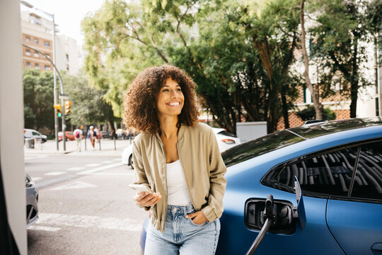 A woman is smiling as she stands next to her electric car at a charging station in a city environment. The scene captures modern transportation and eco-friendly living. - Powered by Adobe