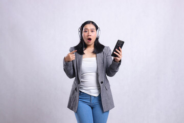 beautiful young office female asia shocked wearing headphone and holding mobile phone while pointing wearing suit isolated on white background. lifestyle, education, entertainment, technology concept