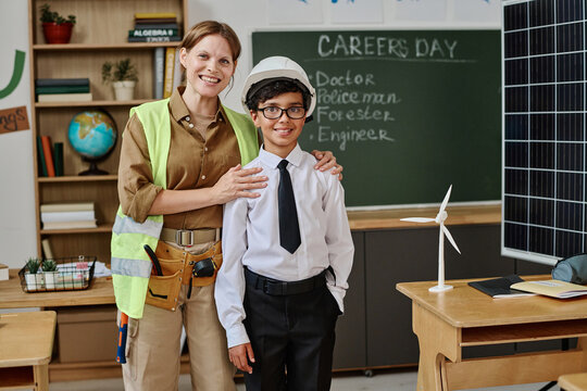 Cheerful female Caucasian engineer and biracial teen student wearing hardhat posing for camera in classroom on career day at school