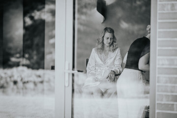  Black and white photo of a woman in a robe, seated and deep in thought, seen through a glass door,...