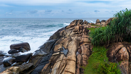 Scenic rocky coastline of Sri Lanka during the day. Waves crash against rugged rocks along the Sri Lankan coastline, surrounded by lush greenery and a serene ocean view under a cloudy sky.