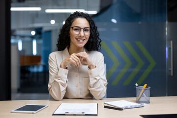 Confident businesswoman wearing glasses smiling at desk with documents, tablet, and stationery in...