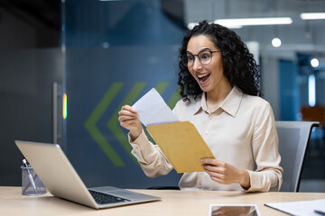 Businesswoman at desk receives good news in envelope, showing excitement and joy. Seated with laptop and phone in office setting.