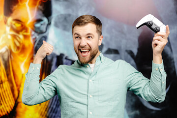 Young bearded man standing at the wall in the room and playing, holding wireless joystick,...