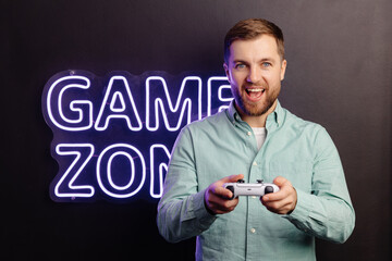 Young bearded man standing at the wall in the room and playing, holding wireless joystick,...