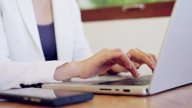 A woman is typing on a laptop computer. She is wearing a white jacket and a blue top. The laptop is on a wooden table