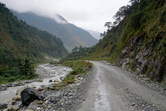 A winding gravel road alongside a serene river in a misty mountain landscape