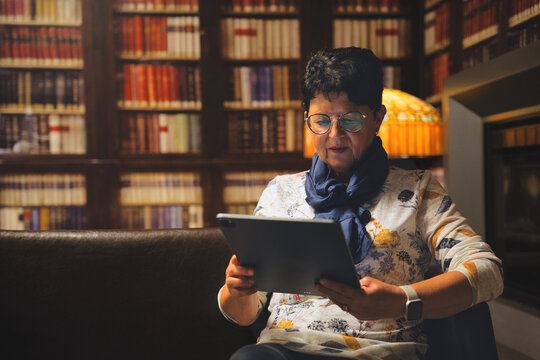 Senior woman is using a digital tablet in a library, surrounded by bookshelves - Powered by Adobe