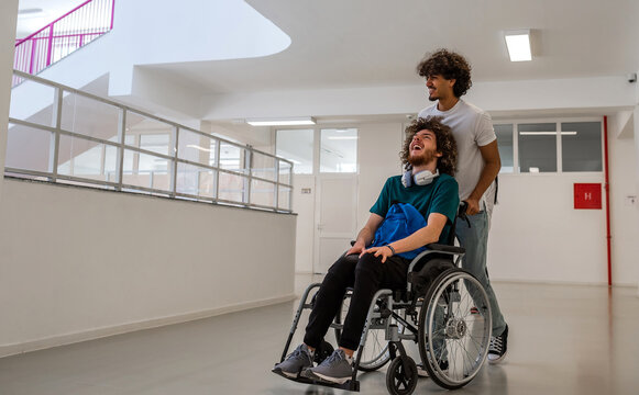 A happy diverse students in university hallway, a classmate pushing a friend in a wheelchair as they walk down the school corridor or male patient smiling with friendly caregiver in hospital corridor. - Powered by Adobe