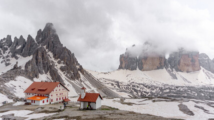 Striking, rocky mountain formation of Tre Cime di Lavaredo partially covered in clouds, with snow surrounding the base and a big mountain refugee with orange rooftop
