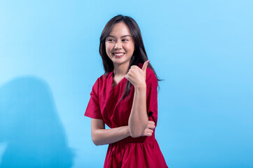 An Asian woman in a red dress smiles confidently while giving a thumbs-up gesture, standing against...