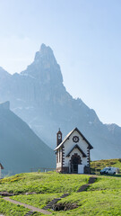 Alpine church on a green field with a rugged mountain peak in the background on a sunny day in Passo Rolle, Dolomites