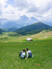 Two hikers resting in a stunning alpine landscape with green meadows and distant mountains under a partly cloudy sky
