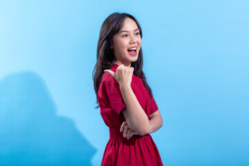 An Asian woman in a red dress smiles confidently while giving a thumbs-up gesture, standing against...