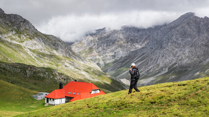 Hiker on a grassy hillside with a red-roofed mountain lodge and rugged, cloud-covered peaks in the background. Ideal for themes of adventure, nature, and mountain exploration