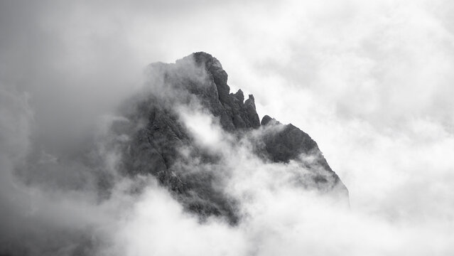 A dark, jagged mountain peak shrouded in thick clouds and mist. The scene is dramatic and atmospheric, with the mountain barely visible through the foggy cover, creating a sense of mystery