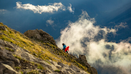 two climbers ascending a steep, rocky mountainside, surrounded by clouds and mist. The sky is dark and dramatic, with rays of sunlight piercing through, illuminating the climbers and the slope