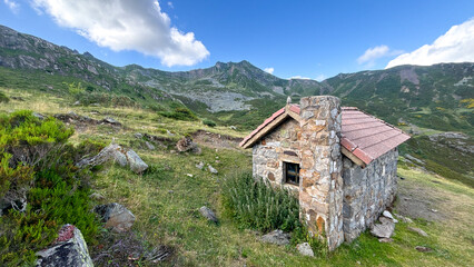 A small rustic stone cabin sits alone in a picturesque mountain landscape, surrounded by grassy hills and rocky terrain under a bright blue sky. Ideal for rural living, solitude, and nature-themed ima