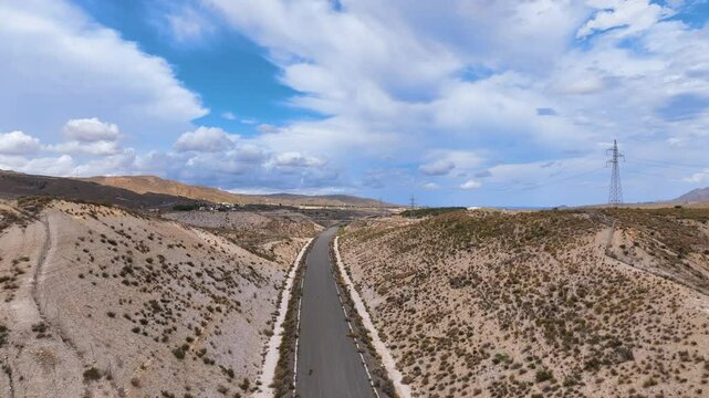 Aerial glides over an emblematic landscape of tussock spotted hills and a long grey vacant road, slowly passing electricity pylons Andalusia Spain.