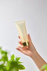 A hand delicately holds a mockup of a white tube with a golden lid against a blurred white background surrounded by lush green leaves, creating a sense of natural beauty and freshness for products.